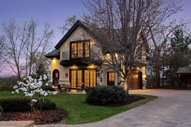 tudor home featuring a front yard, decorative driveway, a balcony, stone siding, and stucco siding