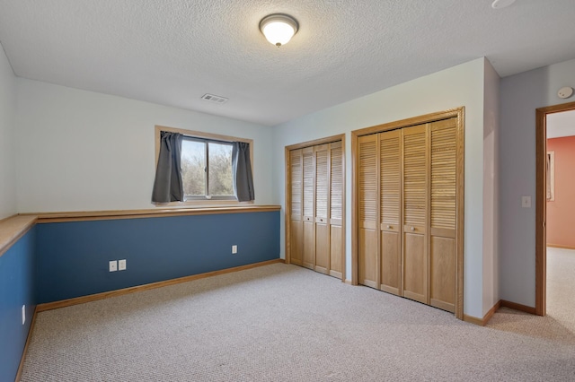 unfurnished bedroom featuring a textured ceiling, carpet, two closets, baseboards, and visible vents