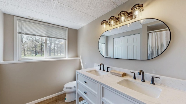 full bath featuring a drop ceiling, toilet, double vanity, baseboards, and wood finished floors