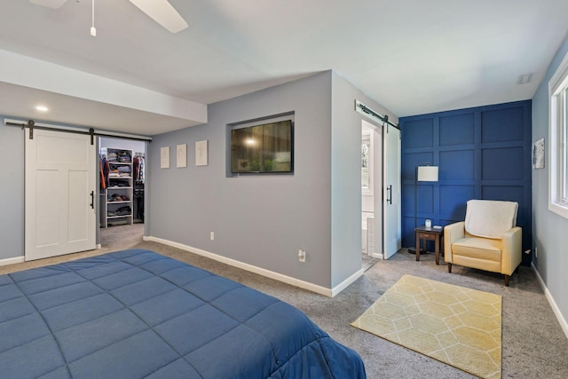 carpeted bedroom with a walk in closet, a decorative wall, a barn door, baseboards, and ceiling fan