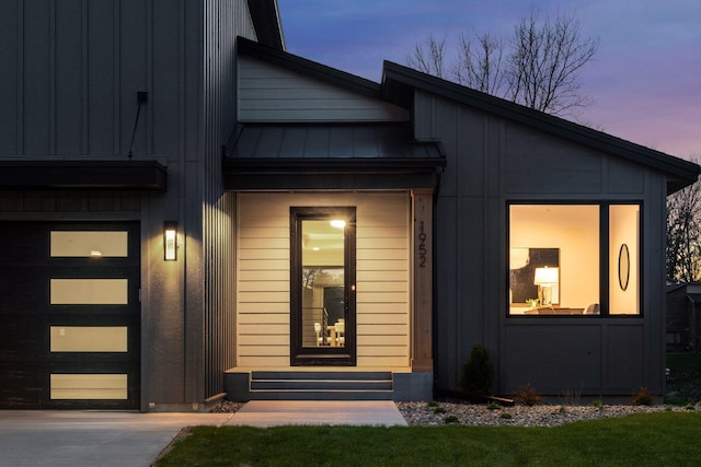 exterior entry at dusk featuring a garage, board and batten siding, concrete driveway, a standing seam roof, and metal roof
