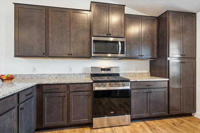 kitchen with appliances with stainless steel finishes, dark brown cabinets, light wood-style floors, light stone counters, and vaulted ceiling
