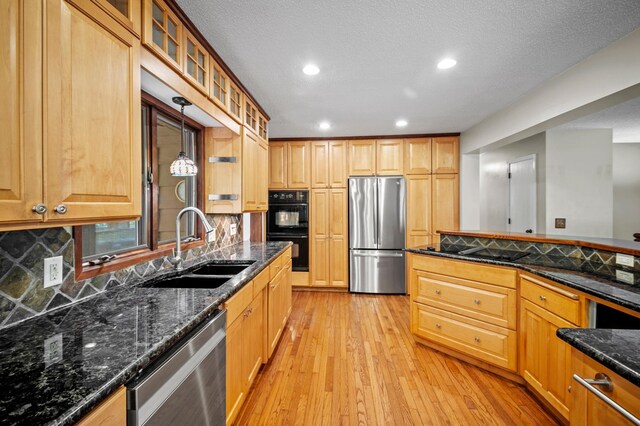 kitchen featuring appliances with stainless steel finishes, a sink, light wood-style floors, decorative backsplash, and dark stone counters
