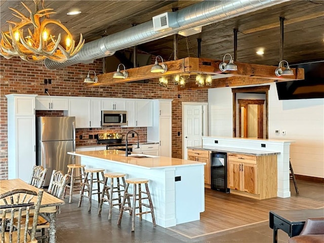 kitchen featuring brick wall, appliances with stainless steel finishes, a sink, beverage cooler, and an island with sink