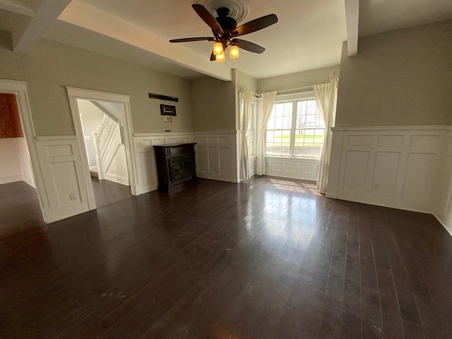 unfurnished living room with dark wood-style flooring, a decorative wall, a fireplace, ceiling fan, and a wainscoted wall