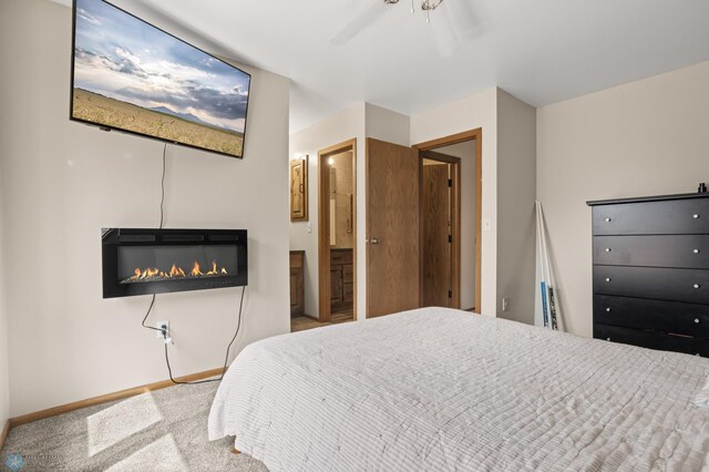 carpeted bedroom with baseboards, a glass covered fireplace, a ceiling fan, and ensuite bathroom