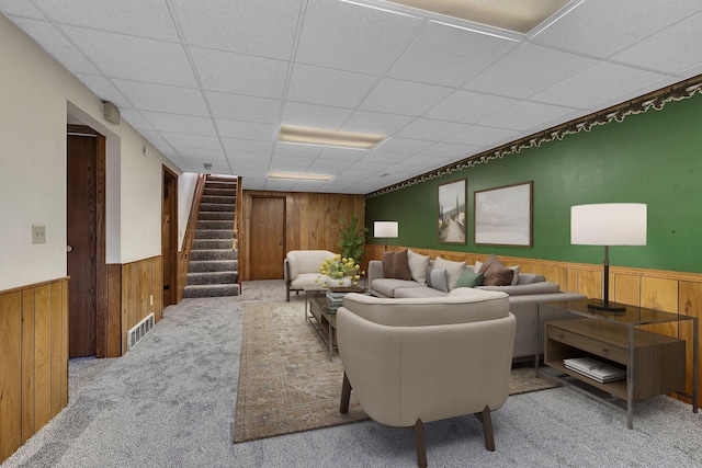 carpeted living area featuring wooden walls, stairs, wainscoting, and a paneled ceiling