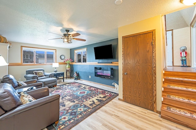 living room with a textured ceiling, a glass covered fireplace, stairs, a ceiling fan, and wood finished floors