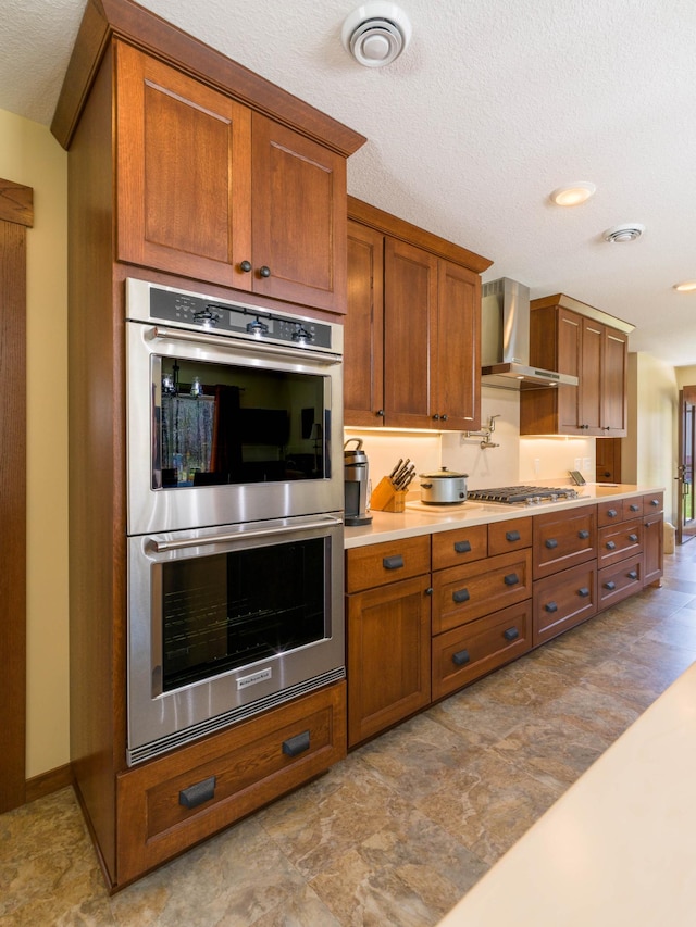 kitchen featuring appliances with stainless steel finishes, wall chimney range hood, brown cabinets, light countertops, and a textured ceiling