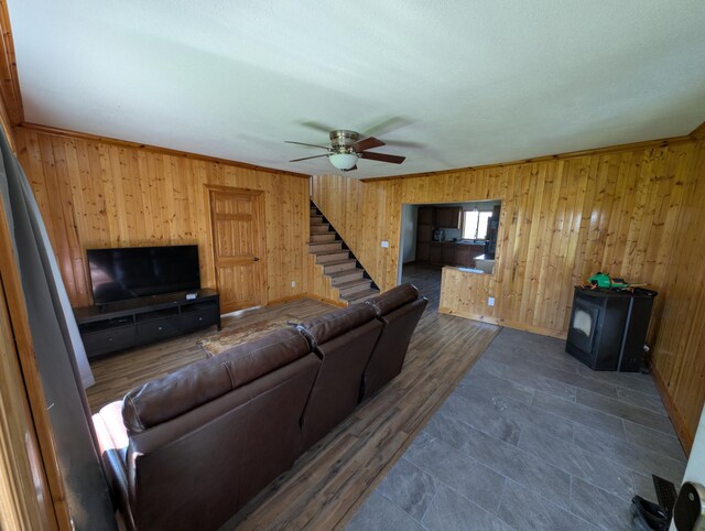 living room featuring wooden walls, stairs, a ceiling fan, baseboards, and a wood stove
