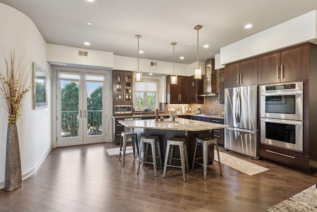 kitchen with dark brown cabinetry, wall chimney exhaust hood, stainless steel appliances, dark wood-type flooring, and a center island with sink