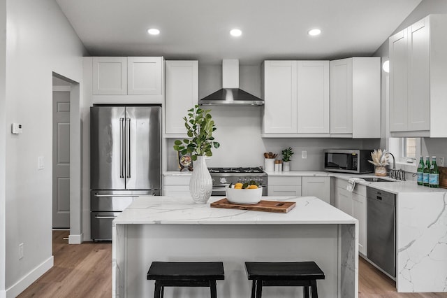 kitchen with wall chimney exhaust hood, high end appliances, a sink, light wood-style flooring, and a breakfast bar