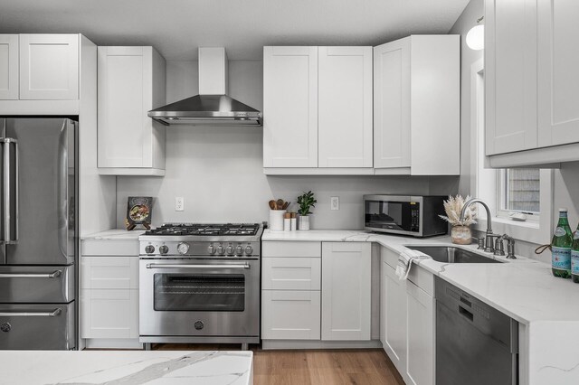 kitchen featuring appliances with stainless steel finishes, wall chimney range hood, a sink, light stone countertops, and light wood-style floors