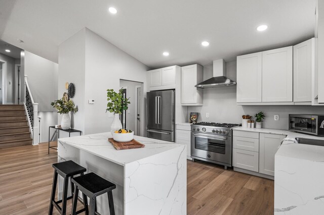 kitchen with high quality appliances, wall chimney range hood, light wood-type flooring, a kitchen bar, and recessed lighting