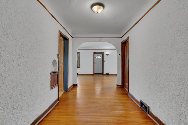 hallway featuring arched walkways, a textured wall, light wood-type flooring, and baseboards