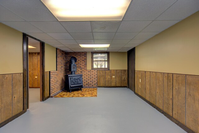 basement featuring wainscoting, a wood stove, wood walls, and a drop ceiling