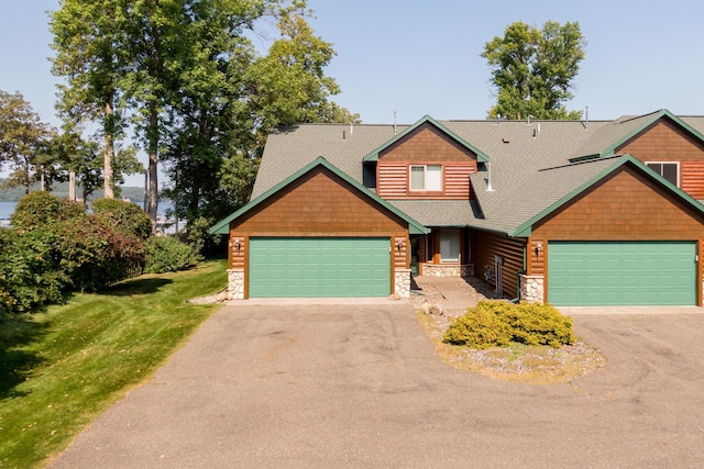 craftsman house featuring stone siding, driveway, an attached garage, a front yard, and roof with shingles