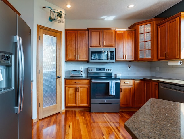 kitchen featuring appliances with stainless steel finishes, light wood-style flooring, dark stone countertops, glass insert cabinets, and brown cabinetry