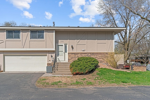 bi-level home featuring brick siding, an attached garage, central air condition unit, stucco siding, and aphalt driveway