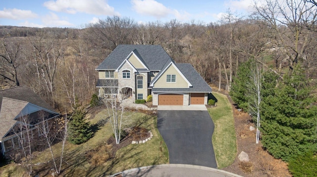 traditional-style home featuring a front lawn, a forest view, driveway, and roof with shingles