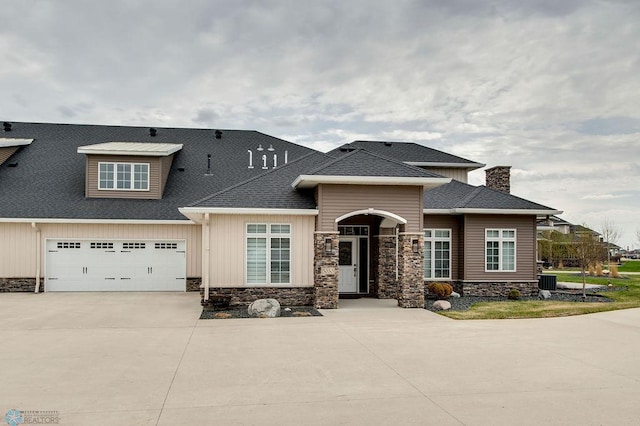 view of front of home featuring roof with shingles, stone siding, driveway, a chimney, and central AC unit