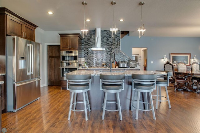 kitchen with stainless steel appliances, dark wood-type flooring, wall chimney exhaust hood, an island with sink, and backsplash