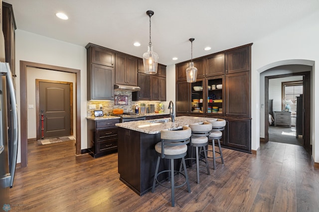 kitchen with arched walkways, under cabinet range hood, dark brown cabinetry, recessed lighting, and tasteful backsplash