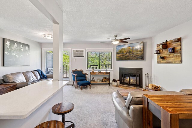 living area with light colored carpet, a glass covered fireplace, a wall mounted air conditioner, a ceiling fan, and a textured ceiling