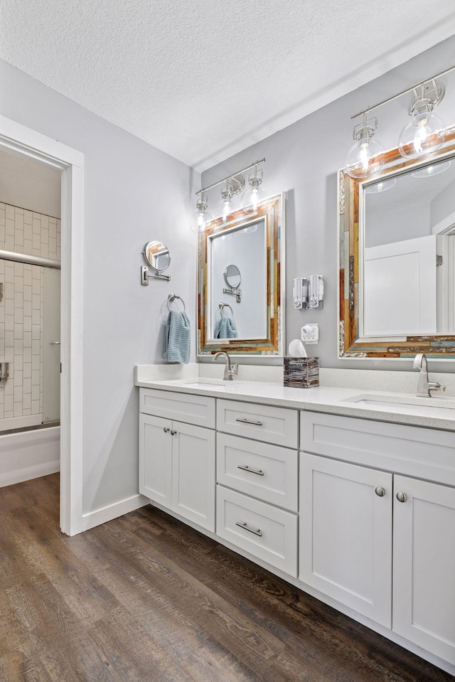 full bath featuring double vanity, wood finished floors, a textured ceiling, and bath / shower combo with glass door