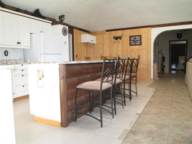 kitchen with wood walls, freestanding refrigerator, white cabinetry, a kitchen bar, and arched walkways