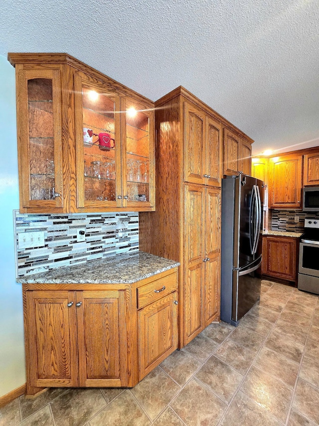kitchen featuring appliances with stainless steel finishes, a textured ceiling, decorative backsplash, brown cabinetry, and glass insert cabinets
