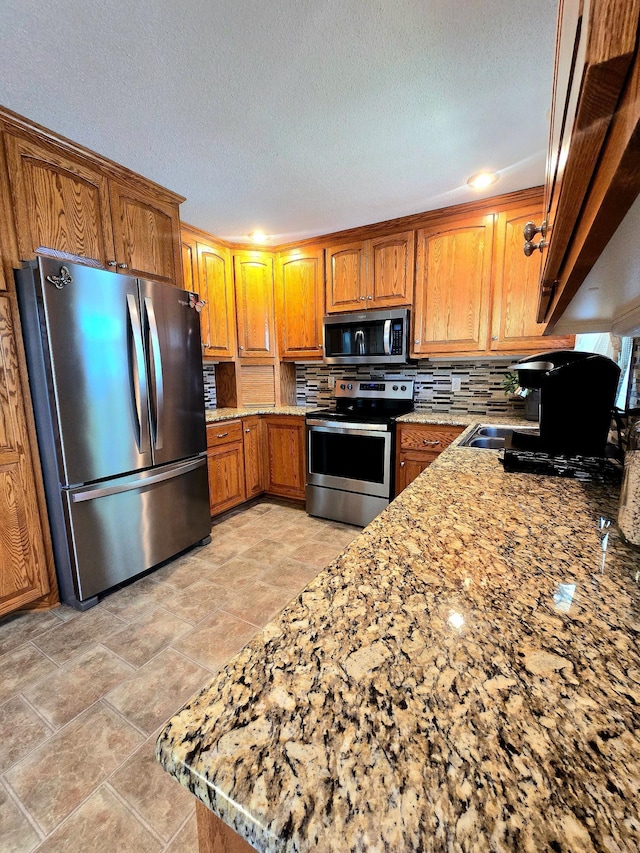 kitchen with stainless steel appliances, brown cabinetry, decorative backsplash, light stone countertops, and a textured ceiling