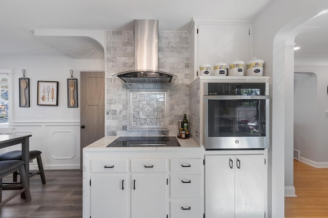 kitchen with stainless steel oven, wall chimney exhaust hood, wood finished floors, arched walkways, and black electric stovetop