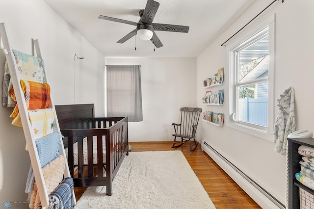 bedroom with baseboard heating, wood finished floors, a crib, and ceiling fan