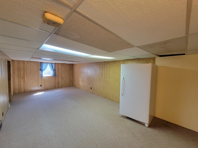 below grade area featuring freestanding refrigerator, a drop ceiling, carpet flooring, and wooden walls