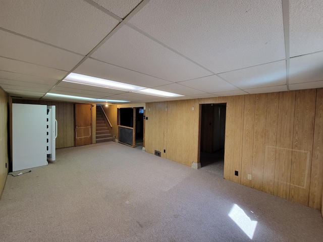 finished basement with a drop ceiling, stairs, light colored carpet, and wood walls