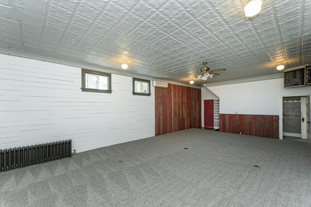basement featuring radiator heating unit, wooden walls, carpet, a wainscoted wall, and a ceiling fan