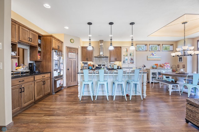 kitchen with stainless steel refrigerator with ice dispenser, wall chimney exhaust hood, brown cabinetry, a breakfast bar area, and a chandelier