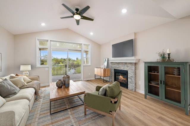 living area featuring vaulted ceiling, light wood finished floors, ceiling fan, a stone fireplace, and baseboards