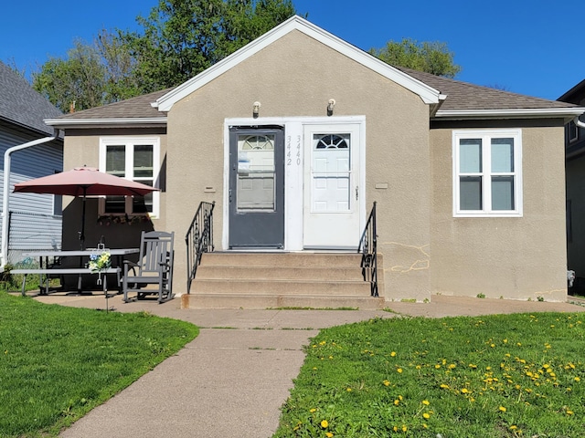 bungalow-style house featuring a front lawn, a shingled roof, stucco siding, and entry steps