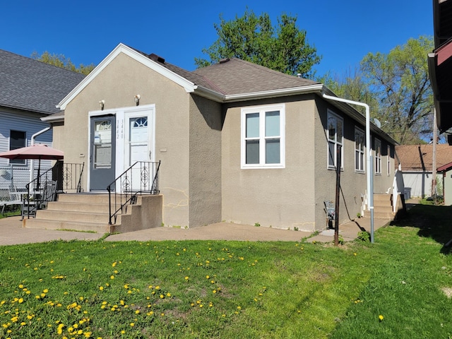 bungalow-style home with roof with shingles, stucco siding, and a front lawn