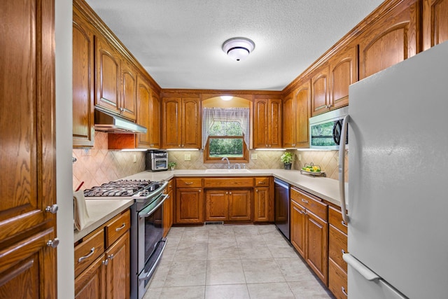 kitchen featuring appliances with stainless steel finishes, under cabinet range hood, brown cabinets, a sink, and light countertops
