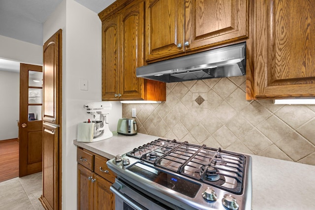 kitchen featuring stainless steel gas stove, ventilation hood, decorative backsplash, brown cabinets, and light countertops