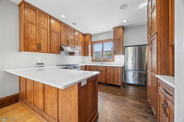 kitchen with appliances with stainless steel finishes, a peninsula, under cabinet range hood, dark wood-style flooring, and brown cabinets