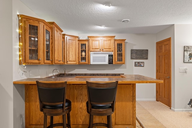 kitchen featuring white microwave, a kitchen breakfast bar, brown cabinets, glass insert cabinets, and a textured ceiling
