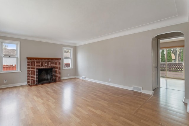 unfurnished living room with healthy amount of natural light, arched walkways, crown molding, light wood-style flooring, and a brick fireplace