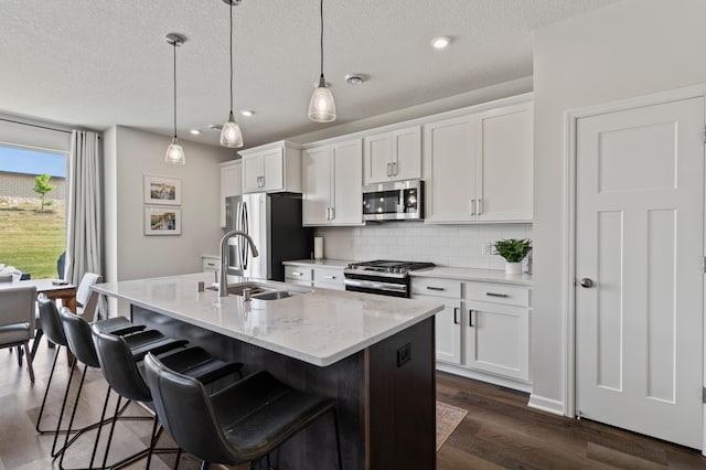 kitchen with stainless steel appliances, an island with sink, a sink, backsplash, and dark wood-type flooring