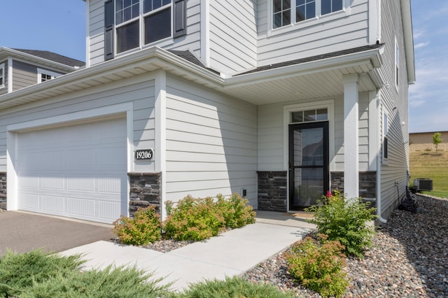 view of exterior entry featuring a garage, stone siding, central air condition unit, and asphalt driveway