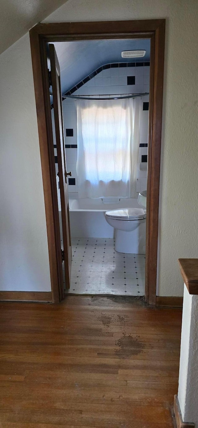 full bathroom featuring wood finished floors, toilet, a textured wall, baseboards, and lofted ceiling