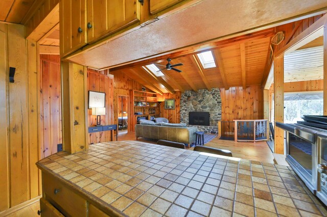 kitchen featuring tile countertops, wooden walls, ceiling fan, a fireplace, and a skylight